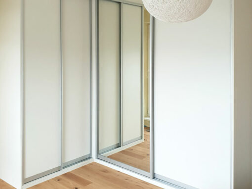 Rohová skříň na míru do ložnice s posuvnými dveřmi v hliníkovím rámu v bílé barvě.