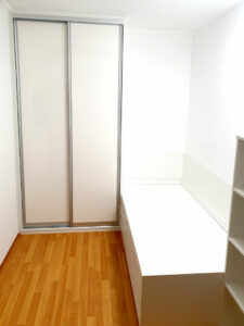 Realizace vestavěné skříně s posuvnými dveřmi s postelí a otevřenou regálem. Vše v bílé barvě.