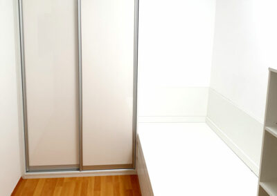 Realizace vestavěné skříně s posuvnými dveřmi s postelí a otevřenou regálem. Vše v bílé barvě.
